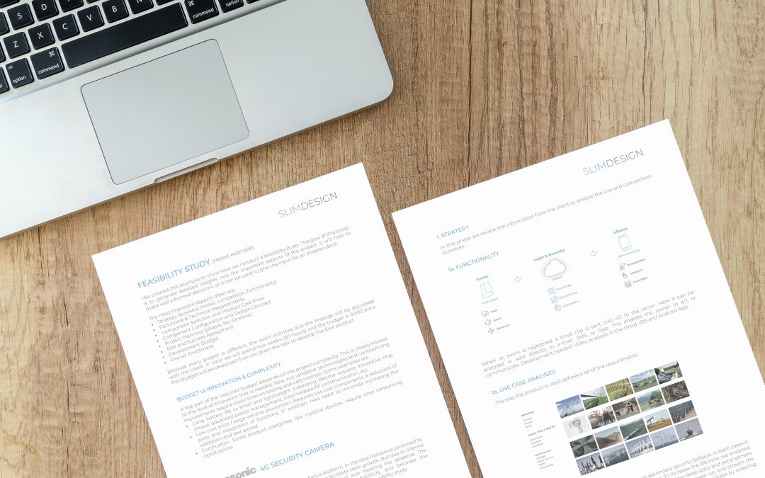 Two printed pages of a feasibility study document placed next to a laptop on a wooden desk. – SLIMDESIGN
