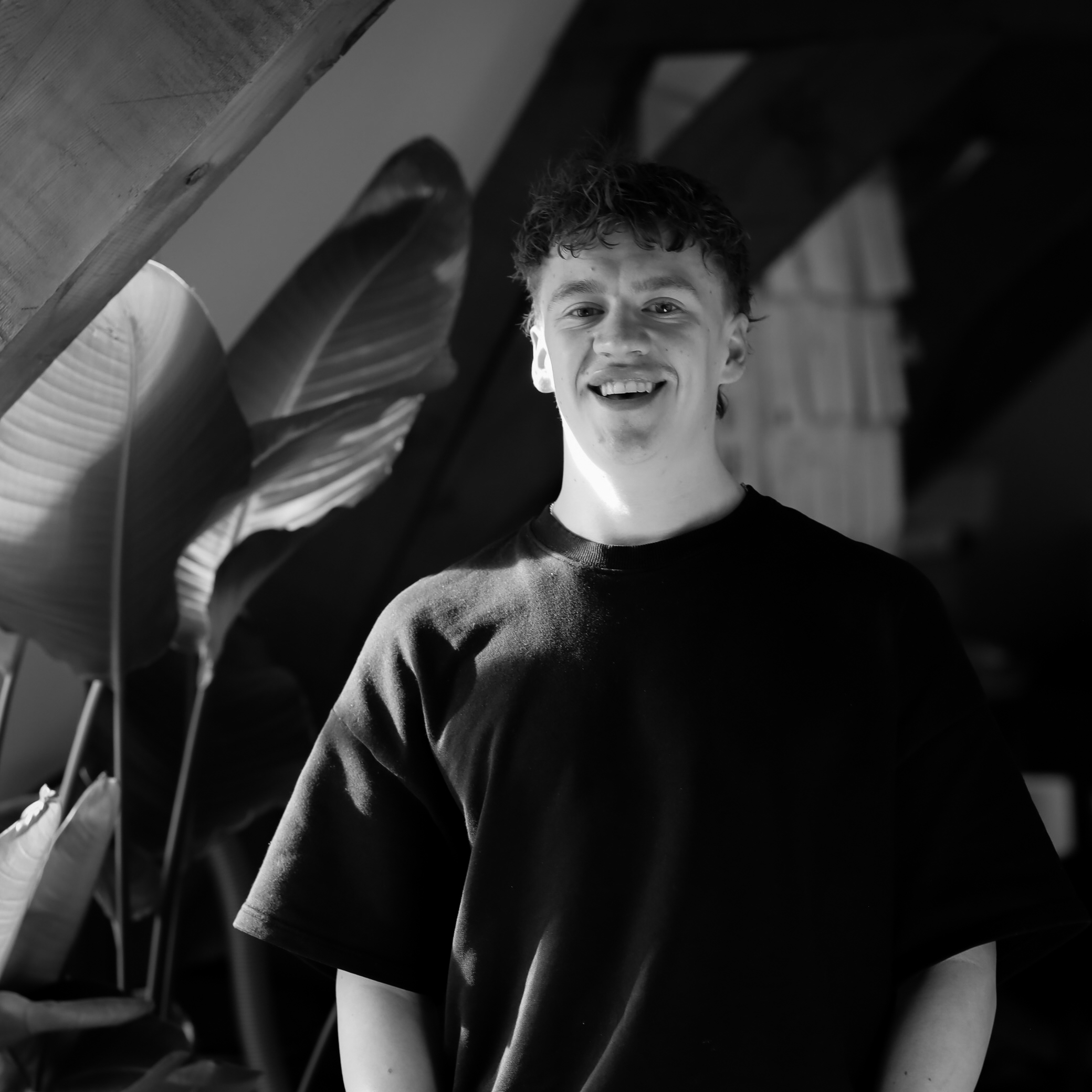 Smiling young man in a black t-shirt standing in front of large green leaves in a monochrome setting.