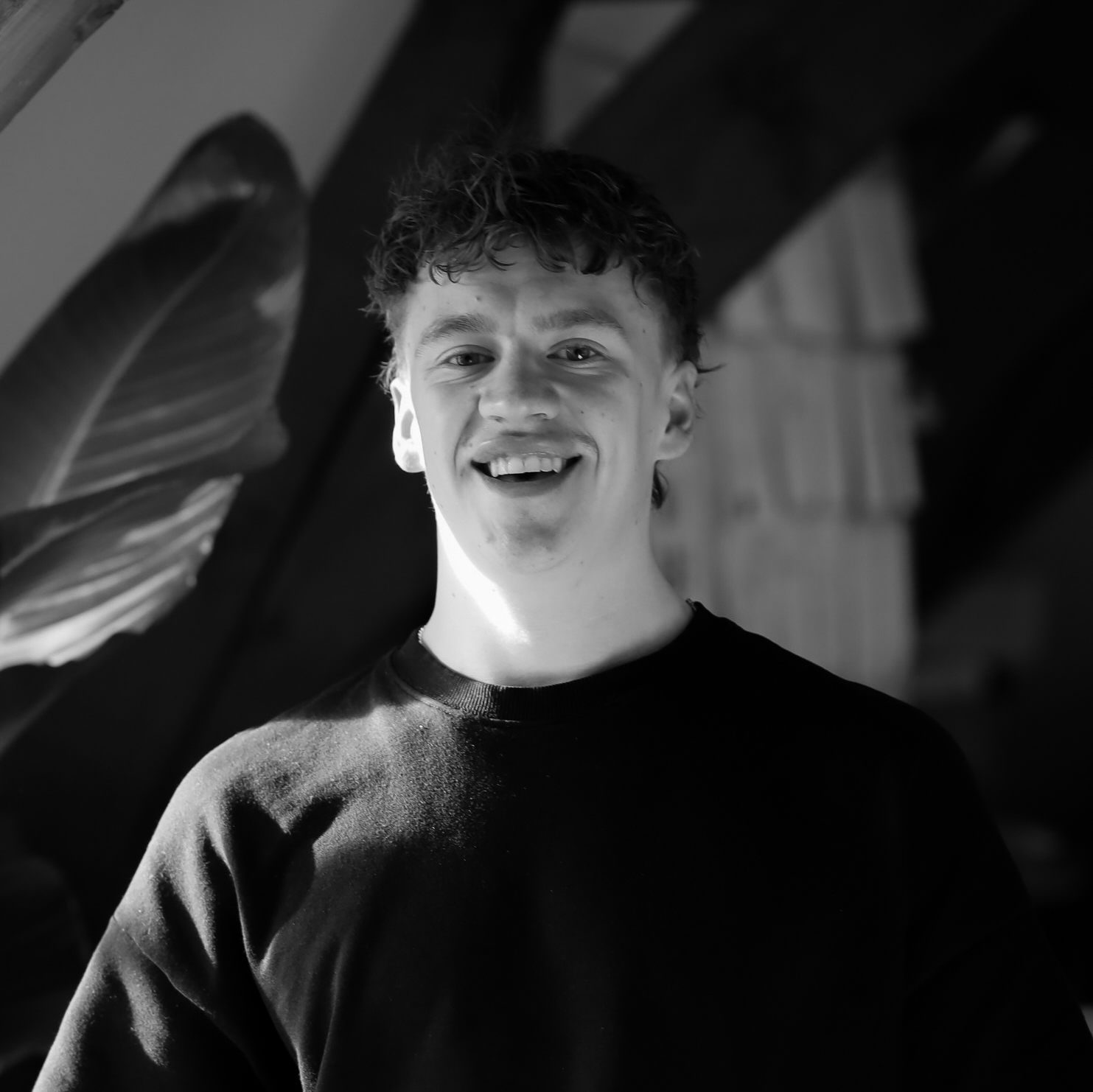 Smiling young man in a black t-shirt standing in front of large green leaves in a monochrome setting.